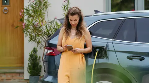 Femme avec un costume sans manches au couleur pêche, regardant son smartphone tout en s'appuyant contre un SUV électrique, avec un chargeur branché.
